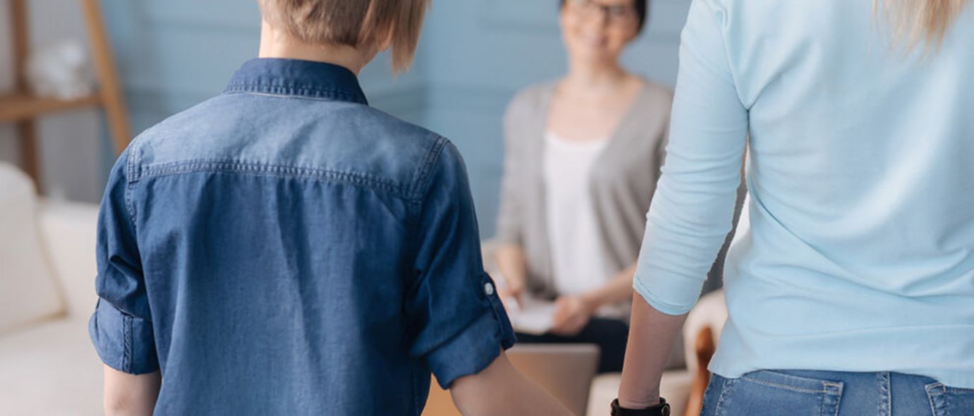 Ein stilvoll gekleideter Junge in Jeanshemd hält die Hand seiner Mutter und posiert mit dem Rücken zur Kamera. Ihm gegenüber sitzt seine Therapeutin