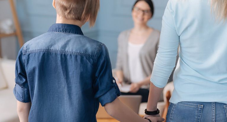 Ein stilvoll gekleideter Junge in Jeanshemd hält die Hand seiner Mutter und posiert mit dem Rücken zur Kamera. Ihm gegenüber sitzt seine Therapeutin