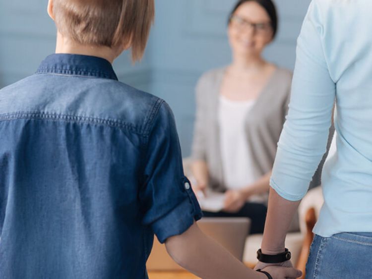 Ein stilvoll gekleideter Junge in Jeanshemd hält die Hand seiner Mutter und posiert mit dem Rücken zur Kamera. Ihm gegenüber sitzt seine Therapeutin