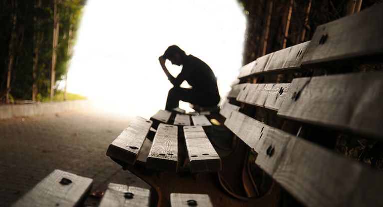 Ein Patient sitzt gedankenverloren auf einer Bank im Wald und hält die Hand an die Stirn